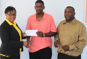 Administrative Officer - Corporate Affairs, Romualda Hyacinth hands over cheque to Castle Bruce Village Councillor, Kurt Cadette while MP Johnson Drigo looks on.”