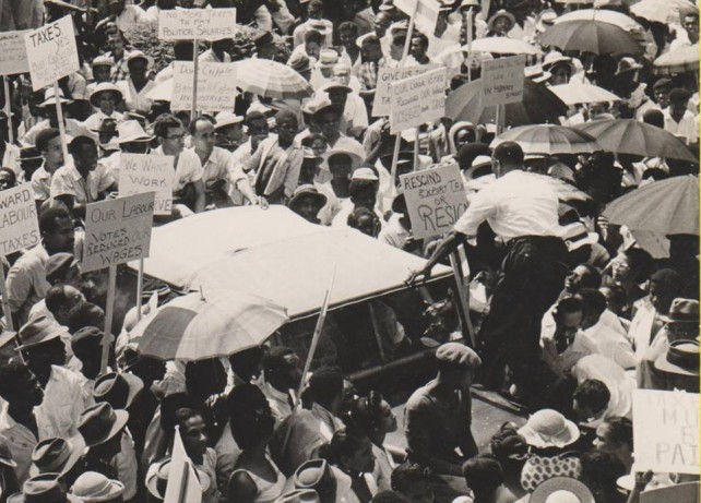 Frank Baron, standing on the Land Rover, backing the camera, leads a demonstration of banana farmers against the ruling Dominica Labour Party government in front of the Government Headquarters on High Street to protest against an export tax on bananas in the early 1960s. Text and photo by Dr. Lennox Honychurch